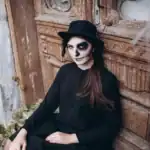21 Stunning Solo Halloween Costumes for Women That Actually Turn Heads (2026) Woman with skull makeup, black hat, and dark outfit for Halloween costume.