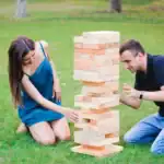 23 Creative Wedding Games for Guests Entertainment Engaged couple playing giant Jenga game outdoors at wedding reception.