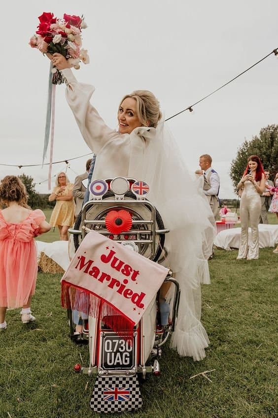 Bride riding a motorcycle with a "Just Married" sign and bouquet, celebrating her wedding reception.