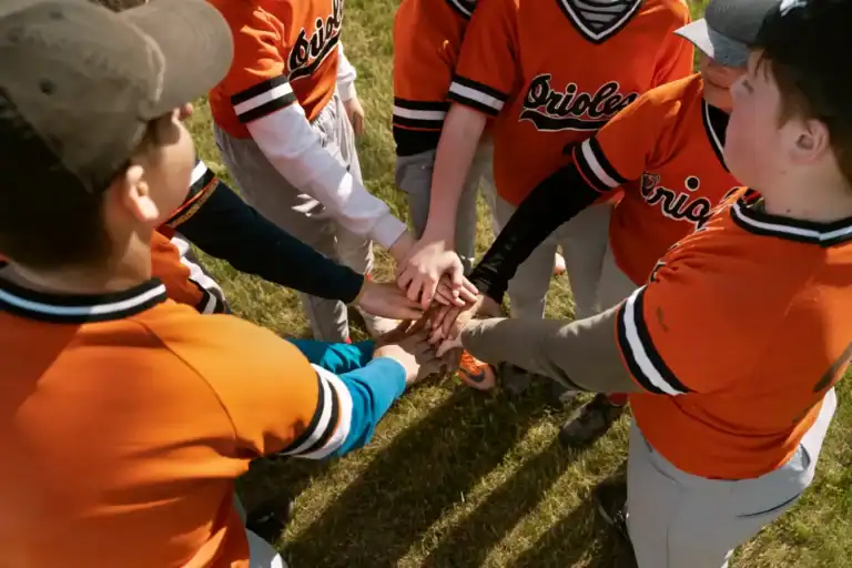 Kids in orange baseball uniforms huddling with hands together on the field.