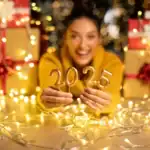 21 Fun Ways to Celebrate New Year’s Eve at Home Happy woman holding "2025" candles with festive decorations and gifts in the background.