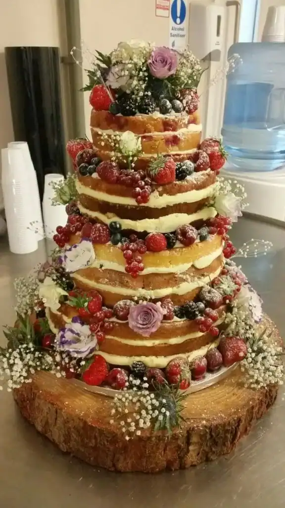 Naked birthday cake showing exposed layers with fresh berries and flowers on top for a rustic outdoor party aesthetic