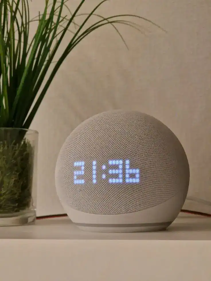 Digital clock with LED display showing 21:36, placed on a white surface next to a potted plant.