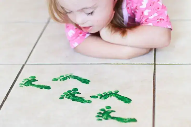 Child creating St. Patrick's Day footprints with green paint on tile floor.