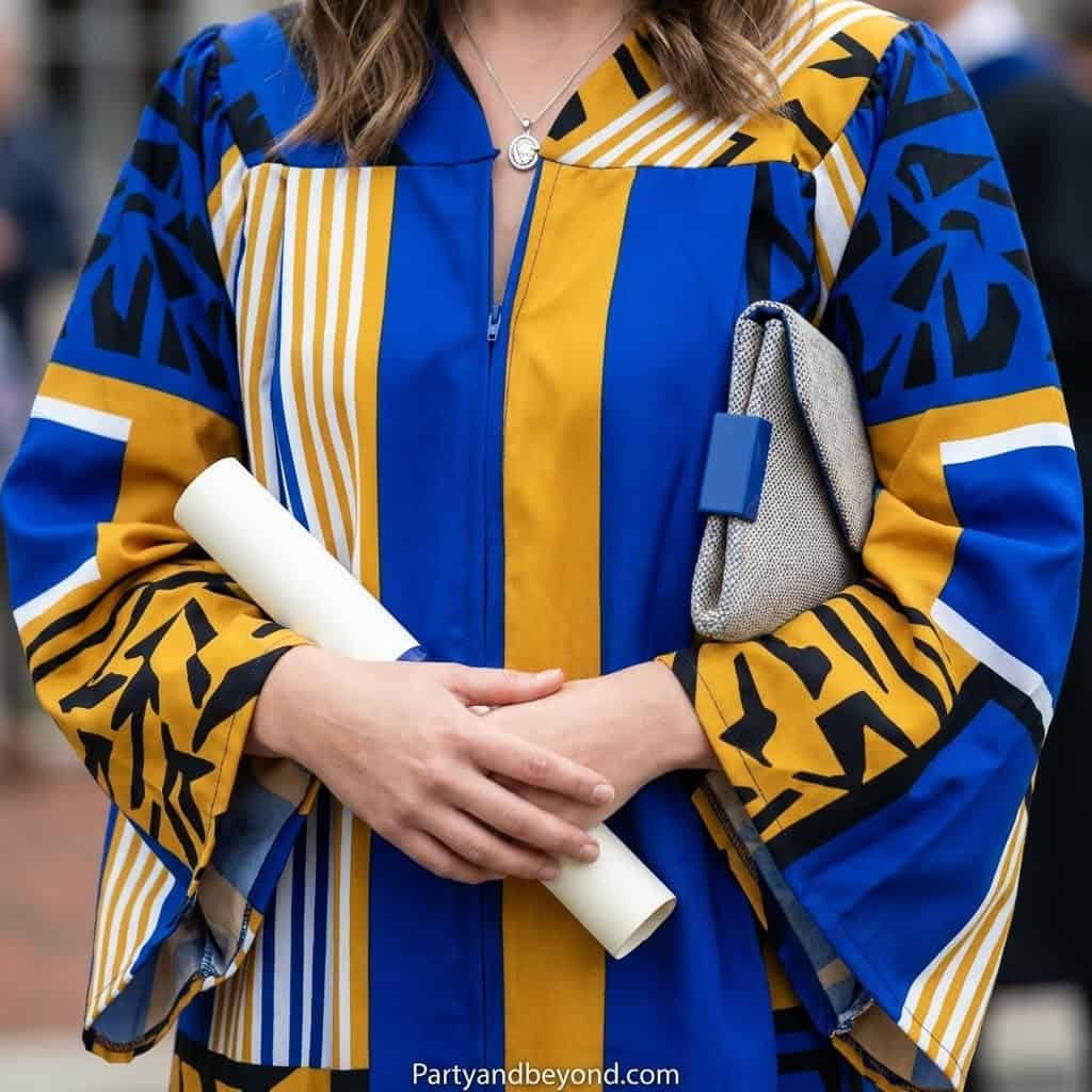 Woman in colorful graduation gown holding diploma and clutch at celebration.