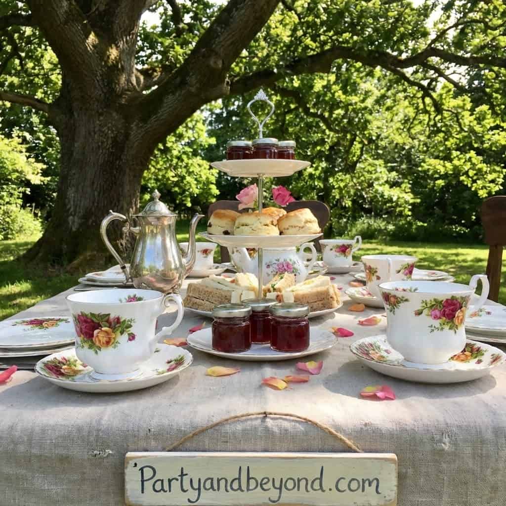 Elegant garden tea party with floral china, tiered cake stand, and jars of jam outdoors.