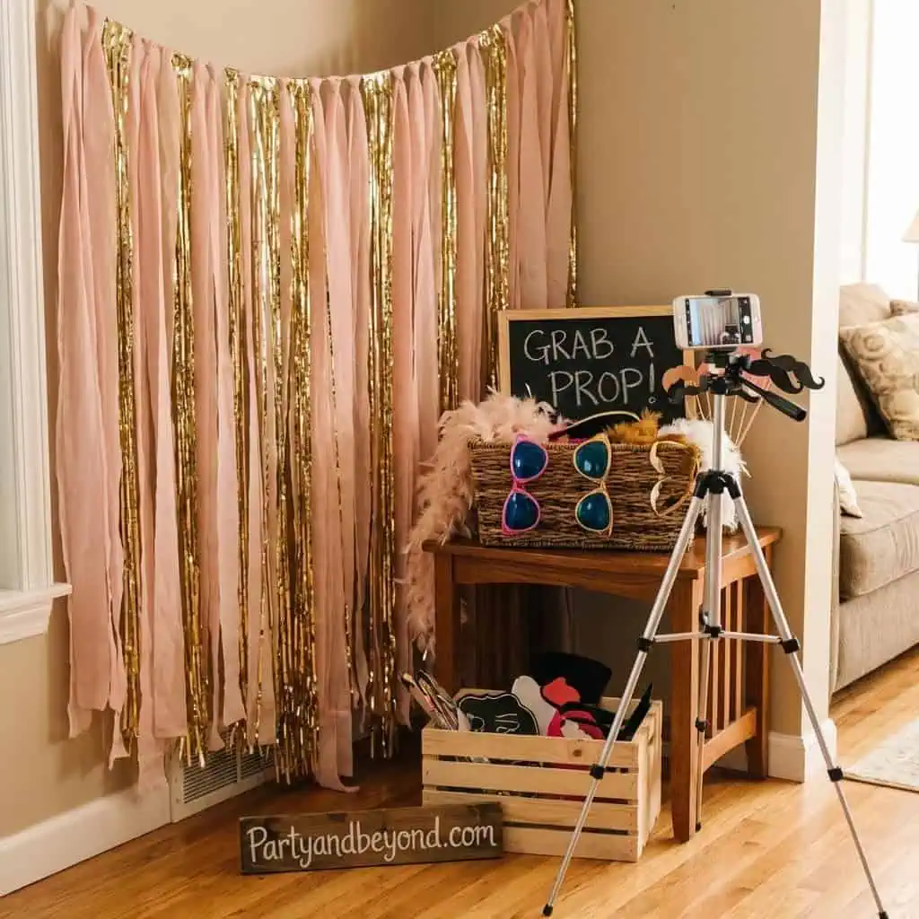DIY photo booth setup with pink and gold streamers, sunglasses, and camera for party fun.