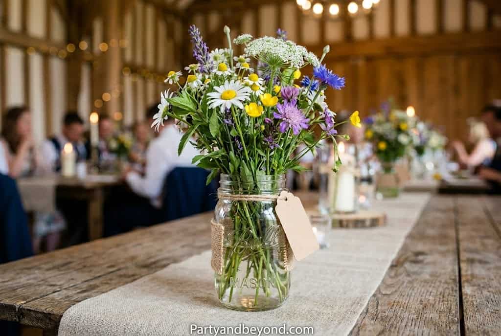 Rustic party table featuring mason jar centerpieces filled with sunflowers, baby's breath, and eucalyptus on a wooden table with soft natural lighting.