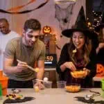 17 Halloween Minute to Win It Games for Adults (Materials & Rules) Halloween party scene where guests in costumes are laughing and playing fast-paced minute-to-win-it games with a timer, creating a fun and energetic group atmosphere.