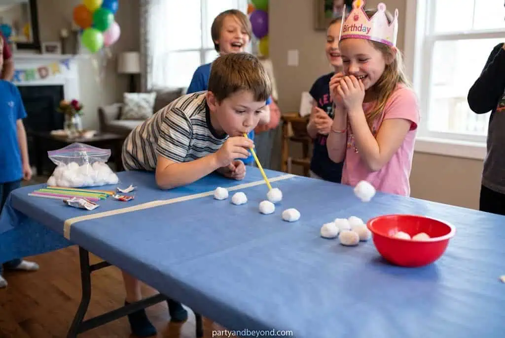 Kids blowing cotton balls across a table with straws during an indoor party game